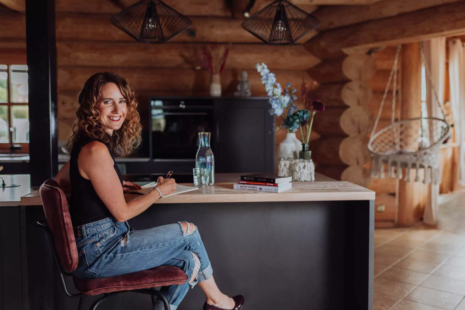 Claudia Burger sitzt in einem Holzhaus seitlich am Tisch und lacht in die Kamera